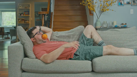 Man Lying on Couch Trying to Get Comfortable with an Orange in His Mouth