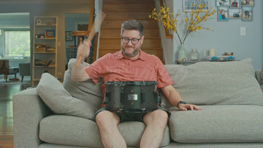 Man on Couch Hits Snare Drum with One Stick Which Confuses His Dog